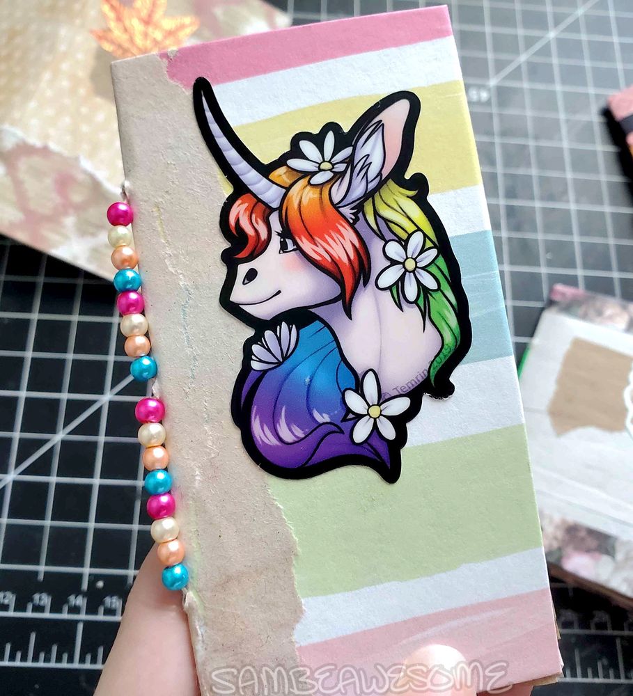 Photo of a long and skinny junk journal with a rainbow paper on the cover and brown craft paper on the spine. The center piece is a rainbow unicorn sticker by Temrin (wildelementstudios), with beads along the hand sewn spine.