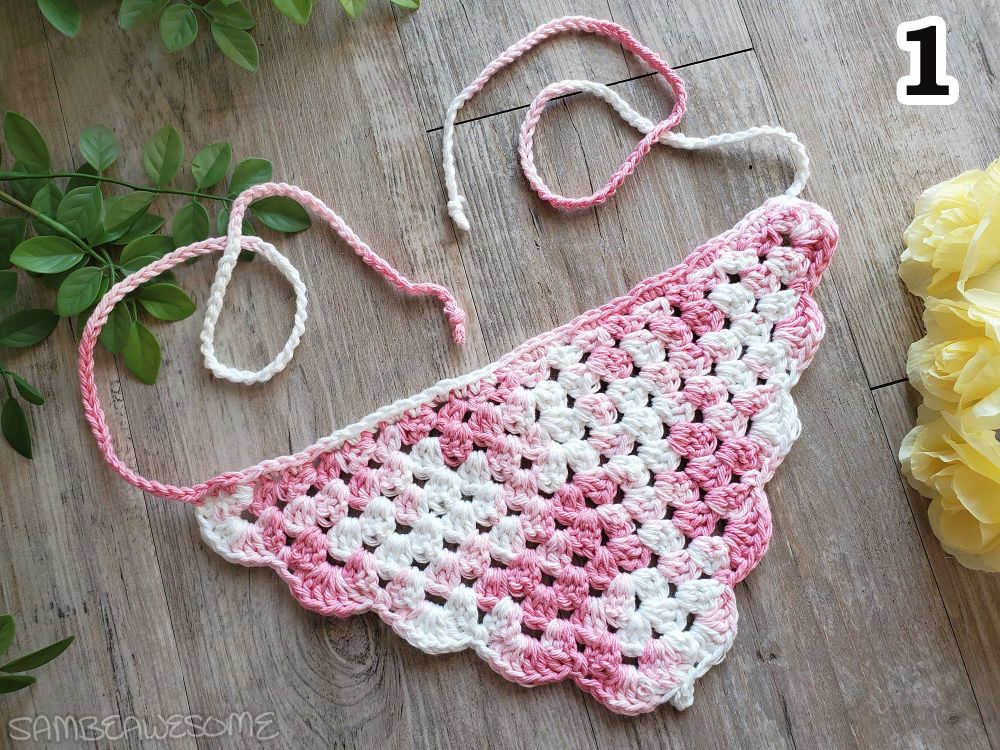 A photo of a handmade crochet bandana that's variegated pink and white, and scallop edge trimming and straps. Fake plants are next to it for the social media photo op.