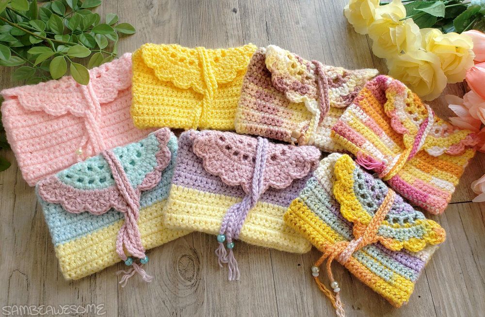 Photo of different handmade crochet pouches with a semi-circle scalloped edge flap, and a yarn chain wrap to close them, with beads on the end of the chain. Each is a different color and in all sorts of color ways, primarily pink and yellow. They are all very bright and pretty.