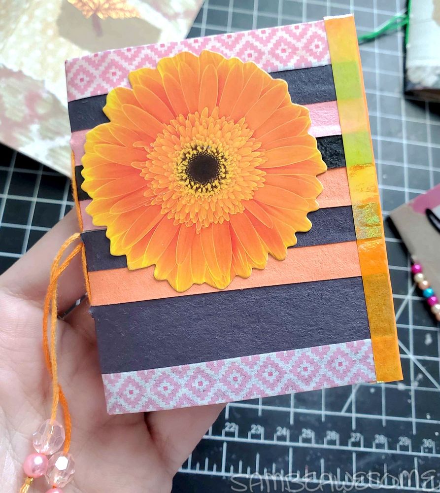 Photo of a small junk journal with various strips of black, pink, and orange craft papers on the cover and washi tapes (pink diamond pattern on the top and bottom, and shiny orange on the ends) on the edges. The cover also has a giant orange flower sticker (purchased from a set from Michaels). Hanging off the excess strands of the hand-sewn spine is various color coordinating beads.