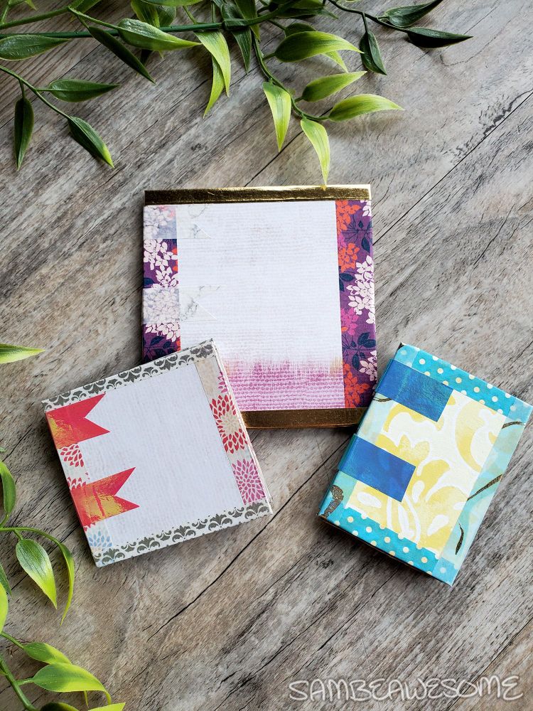 A photo of three junk journals made with fancy craft papers glued over a cardboard cereal box, with washi tape edging, and washi tape binding. One is more purple and gold themed, another is pink and silver themed, and the last is blue and yellow themed.