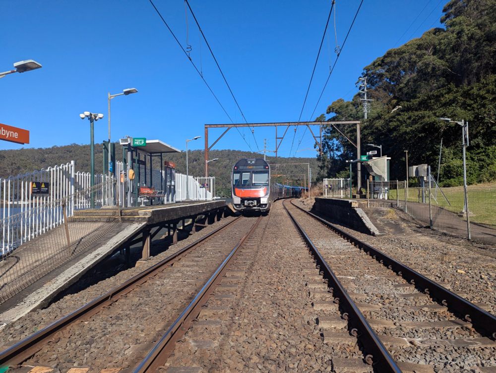 The rear of an orange, silver and black double-decker intercity Mariyung train on the left track, departing the small station of Wondabyne, with some other silver and black carriages visible as it curves further down the tracks. Two tracks with concrete sleepers and gravel ballast are visible, with the right track vacant. Flanking the tracks are two short concrete platforms each with a small shelter and a bench. Overhead catenary wires are visible with a gantry supporting them toward the far end of the platforms. The sky is bright and clear and there are forested hills in the background.