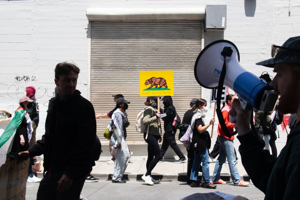 Marchers in front of the ICE building, which is unmarked. In the foreground is a man with a megaphone. Background: A woman holds a sign that says "don't tread on us", depicting the bear from the California flag eating a snake. 