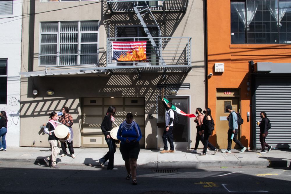 Marchers in front of a building. A upside-down American flag with flames printed on it hangs from a balcony above demonstrators. 
