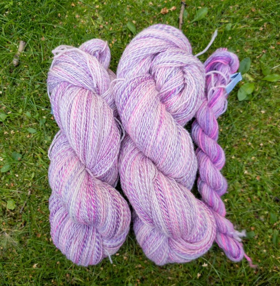Three twisted hanks of yarn on green grass. Two of the hanks are two-ply, one white and one purple-pink thread. The colour changes a lot in the yarn. The third hank is chain plied yarn using only the pink-purple thread. 