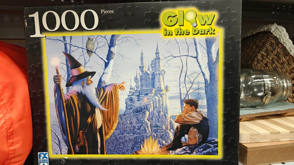 A 1000 piece glow in the dark puzzle depicts a wizard in full regalia pointing toward a white castle atop the edge of a cliff in the distance. A young boy sits near a fire and looks like he is so over this adventure.