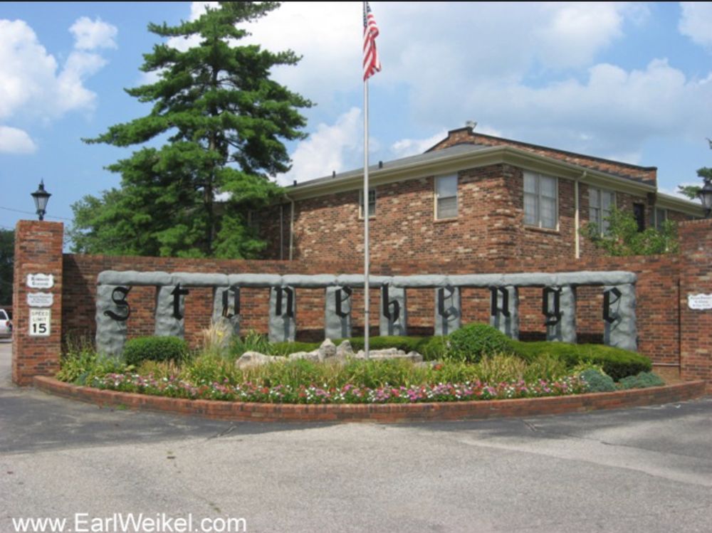 A brick entrance to a set of brick condominiums and in front of the brick wall is a line of 10 grey stone pedestals linked at the top by grey lintels. And on each grey pedestal there's an ornately shaped black letter, together spelling Stonehenge.