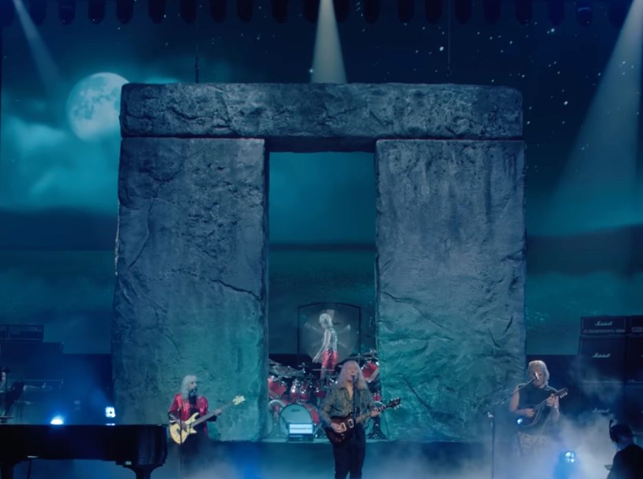 Some rather elderly musicians in front of a very large and square trilithon made of fake stone to look like one from Stonehenge.