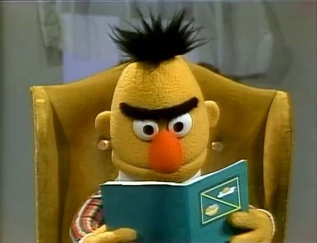 Bert, the muppet, reading a green book while sitting in a yellow chair.