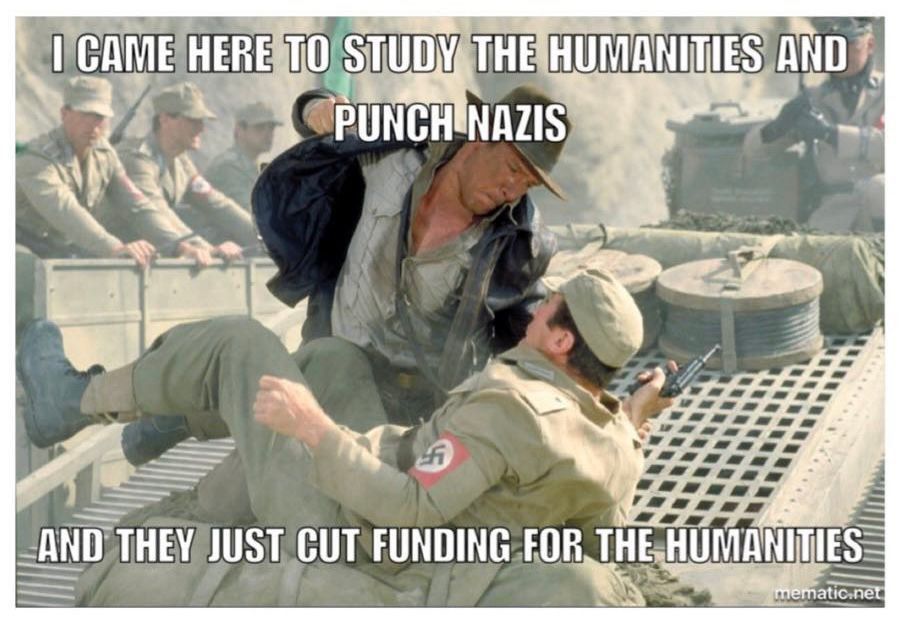 A humorous meme featuring a still from the movie "Raiders of the Lost Ark," showing Indiana Jones punching a Nazi soldier.

The image is split into two sections:

Top Section: A film still from the movie "Raiders of the Lost Ark" shows Harrison Ford as Indiana Jones punching a Nazi soldier. Indiana is in the center of the image, leaning over and punching down at the soldier who is falling backward. The soldier is wearing a tan uniform with a swastika armband.  In the background, other soldiers stand watching.

Bottom Section: White text on a black background reads: "I CAME HERE TO STUDY THE HUMANITIES AND PUNCH NAZIS AND THEY JUST CUT FUNDING FOR THE HUMANITIES."