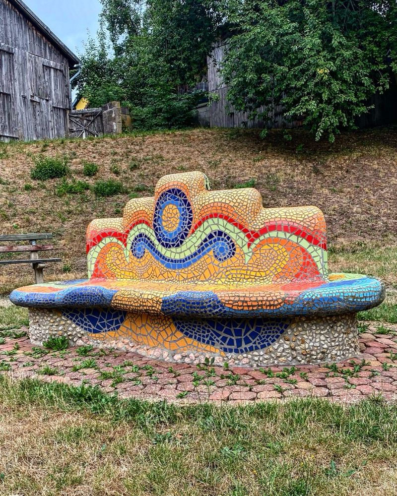 Eine Bank steht auf einer Wiese an einem Hang. Im Hintergrund oben am Hang links eine alte Scheune und rechts große Büsche. Die Bank ist mit bunten Mosaiksteinen gestaltet. Geschwungene Formen in orange, rot, gelb, grün, blau und türkis. Drumherum am Boden rötliche Pflastersteine. 