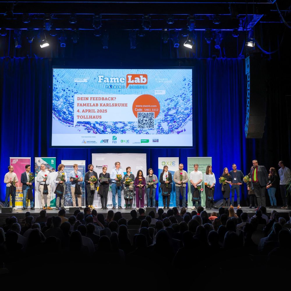 Foto: FameLab Germany / Karlsruhe, Jürgen Rösner Beschreibung: Gruppenfoto beim FameLab Karlsruhe: 12 Nachwuchsforschende und die Jury stehen mit Blumensträußen auf der Bühne des Tollhauses, vor ihnen das Publikum, hinter ihnen eine große Leinwand mit Eventinfos. 