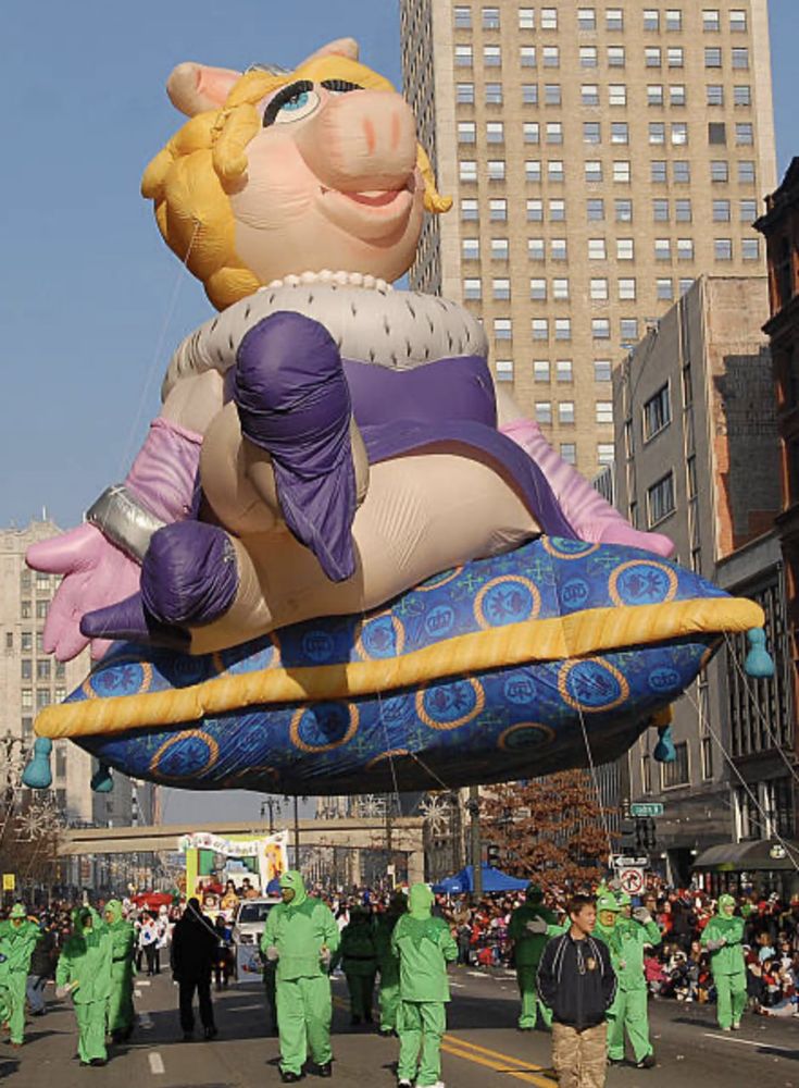 Miss Piggy balloon in a parade, she is lounging on a pillow and her balloon is being handled by people dressed as Kermit