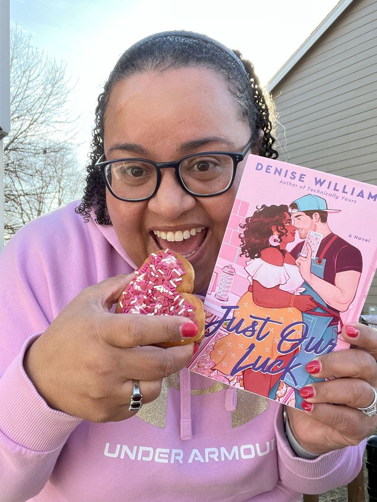 Denise in a pink sweatshirt holding a copy of Just Our Luck and eating a donut with pink and white sprinkles.