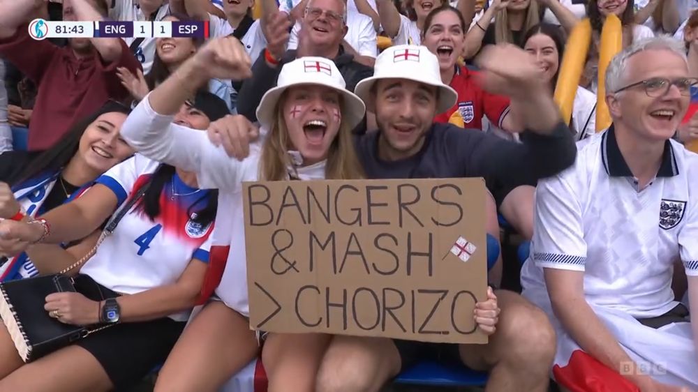Picture of some people in the England crowd at the England vs Spain final of Euro 2025. Two of them are holding up a sign that says “Bangers & Mash > Chorizo”.