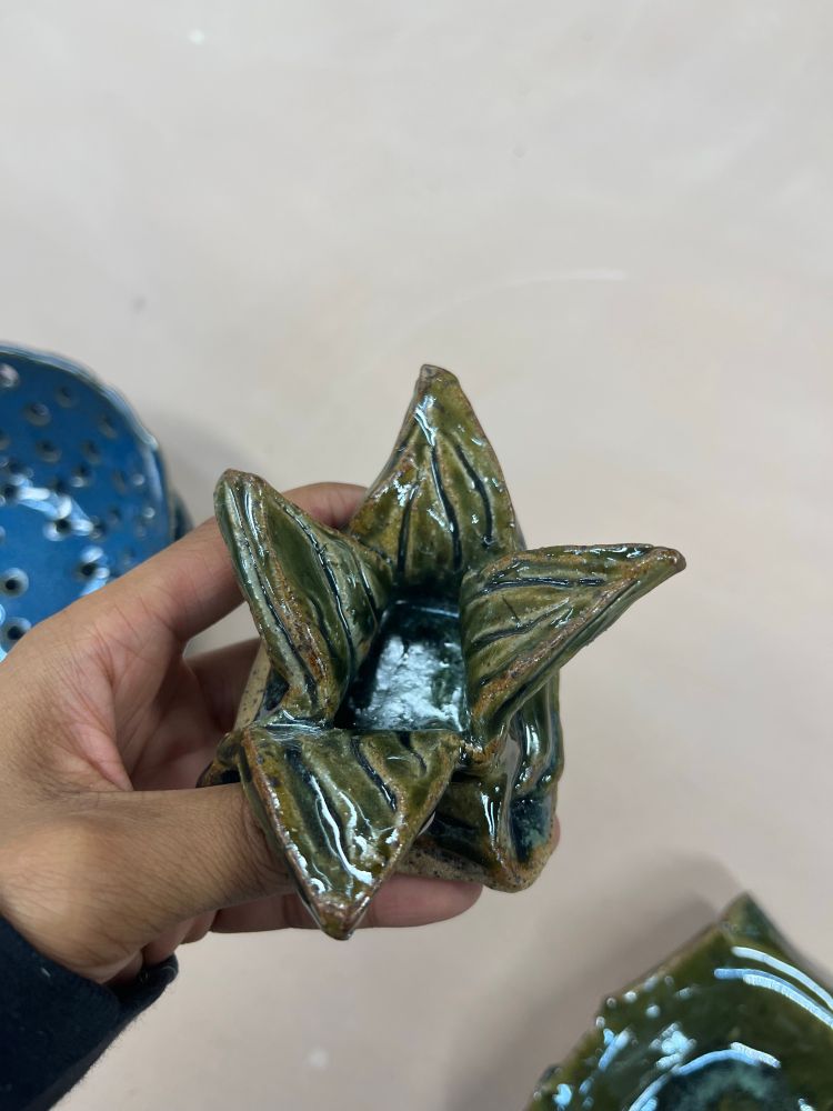 A few of my ceramic pieces sit on the table while a brown hand holds up a green square shaped bowl with four petal like triangles sticking out from the rim. There are various lines in the center making it leaf like. Part of the brown of the clay is showing through adding extra dimension.