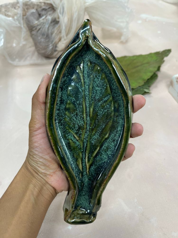 A brown hand holds a finished glazed ceramic piece. It’s in the shape of a leaf and is colored with a dark green. It’s also slightly shaped like a fish. In the background in the table is a dry leaf that crumbled into pieces later. 