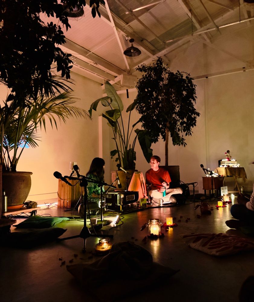 A couple on the fooor, a woman with long dark hair wearing green and a man with short light brown hair and a red top surrounded by instruments, mics, candles and plants 