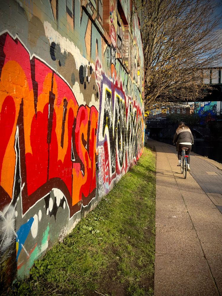 Photo of cyclist riding towards a bridge along regents canal with graffiti on a wall and a grass verge 