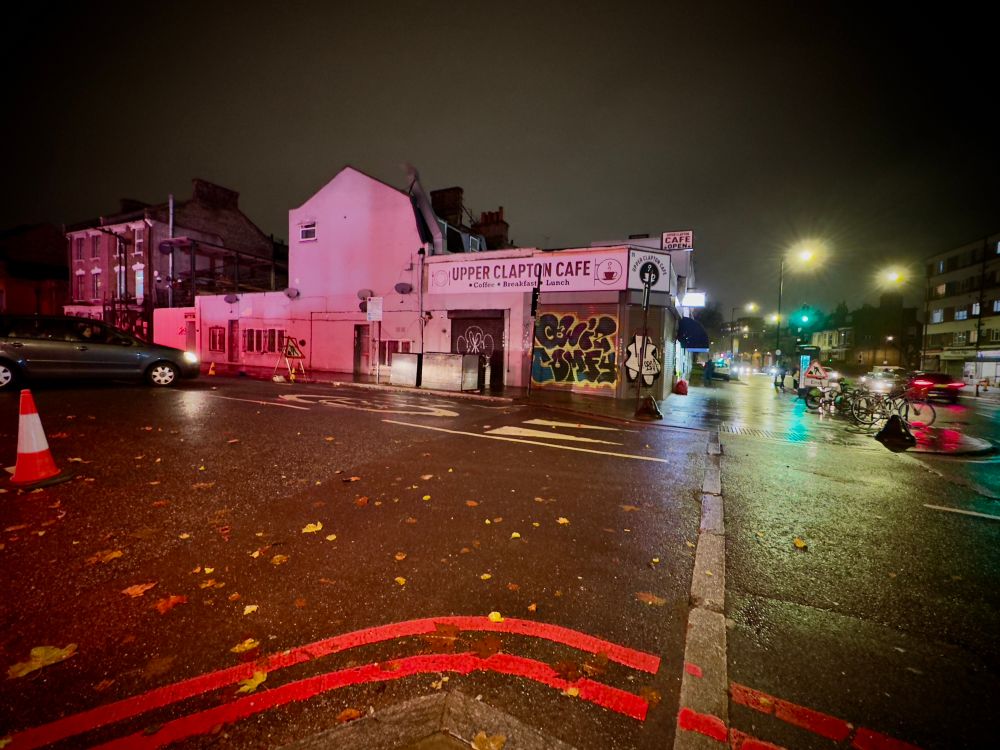 A building looking pink in the lights, red looking road markings, a traffic cone and cars and lights in the rain 