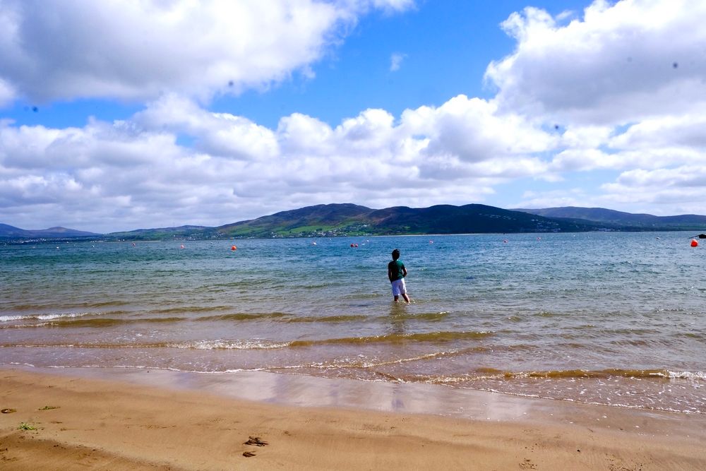 A child stands in the water 
There’s sand in foreground and a headland in distance 
The sky is blue and clouds are fluffy and white 