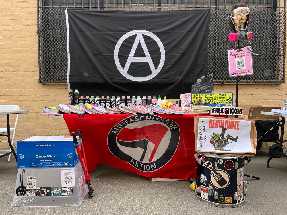 Picture of zine table. Giant anarchist flag behind table. Zines, spray paint, box of free stickers, sit on table. A drum with prints sits in front as well as another box of zines with some bolt cutters. 