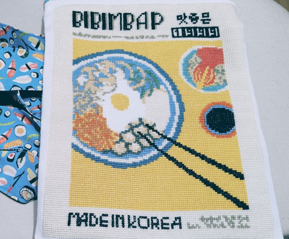 A photo of a cross stitched bag. The front design is of bibimbap Korean food, and the bag has sushi fabric.