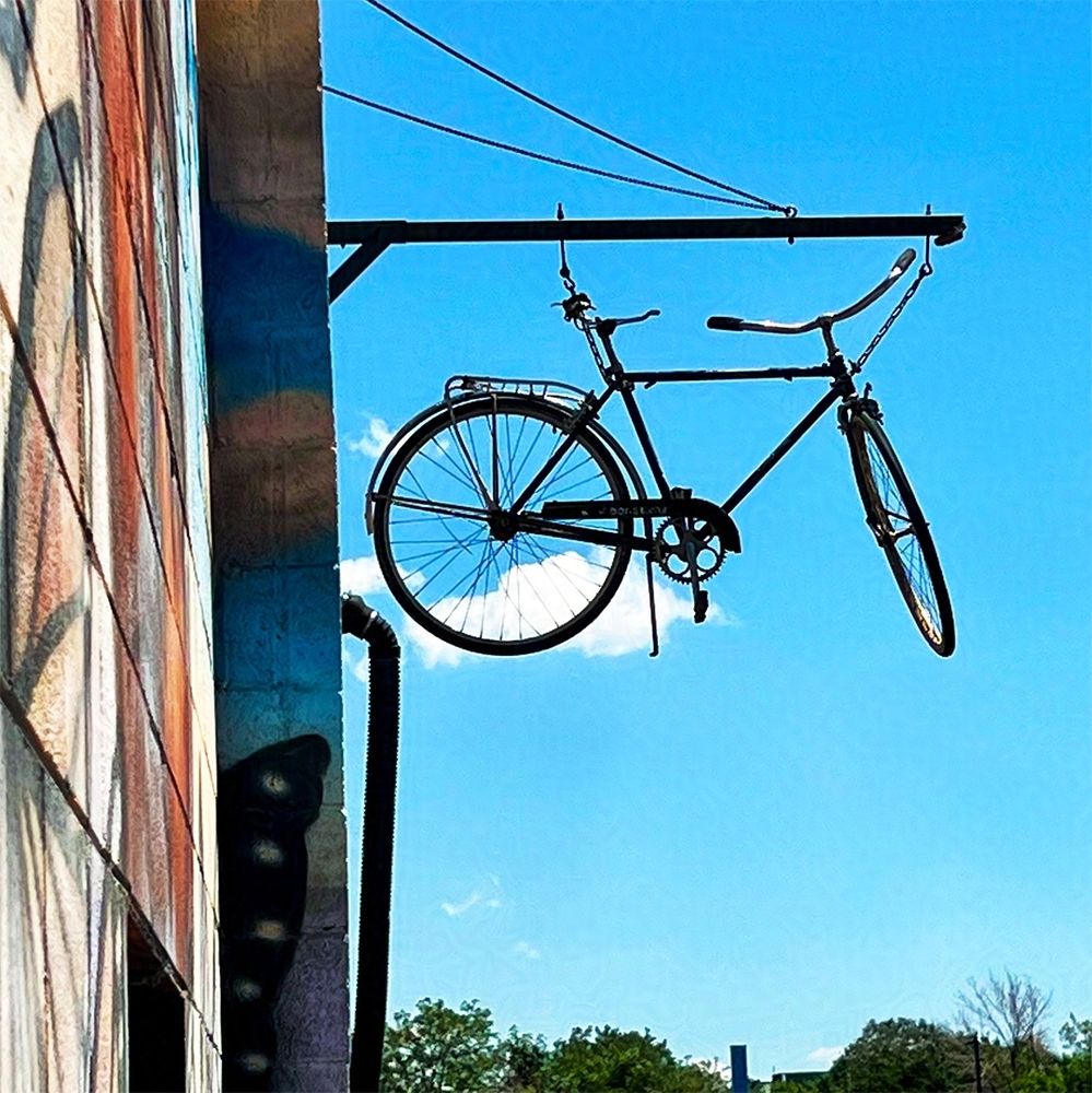 Photo of a bicycle hanging from a second-story pole on the mural-covered Bike Co-op building in Fort Collins, CO. It floats with the clouds in a blue sky.