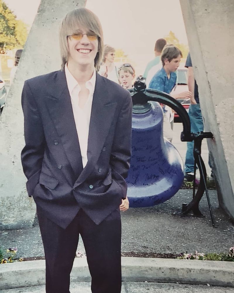 A skinny teenage creature, surprisingly with blonde hair (I was naturally blonde as a kid and it darkened with age). My hair is mid-length and straight, just to my shoulders, I have a huge smile and I am wearing small, oval yellow-tinted glasses. I am wearing a two piece purple suit that is too large for me, with a pink button-up shirt underneath.