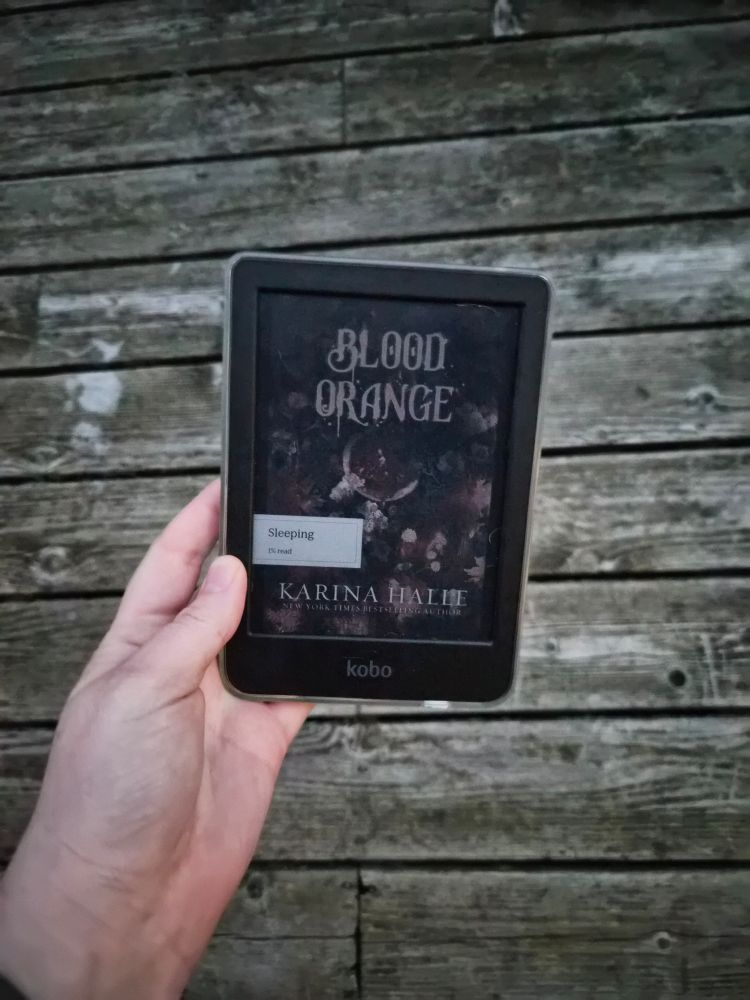A hand holding an ereader displaying the cover for Karina Halle's book,  Blood Orange. Background of photo is a grayish, distressed wood plank wall.