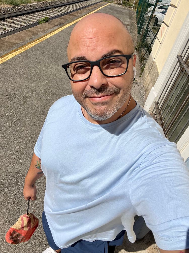A gorgeous male looking person taking a selfie. They have a shaved head, black-framed glasses, a celeste t-shirt and blue shorts with white shoes. The person is smiling at the camera and squinting because the sun is illuminating their face. They’re standing near a singular railroad at a small train station.