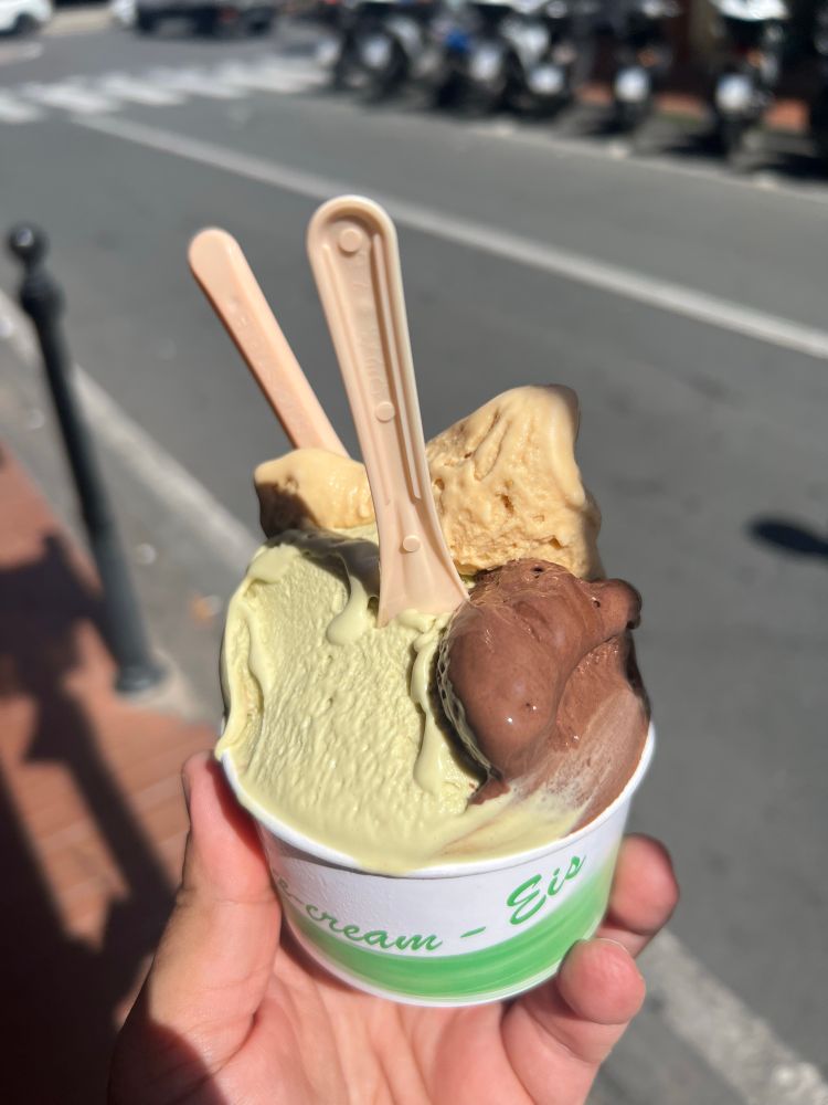 A hand holding a small paper cup with chocolate, pistachio, walnut, and salted caramel gelato. Two pink plastic spoons stick on top.