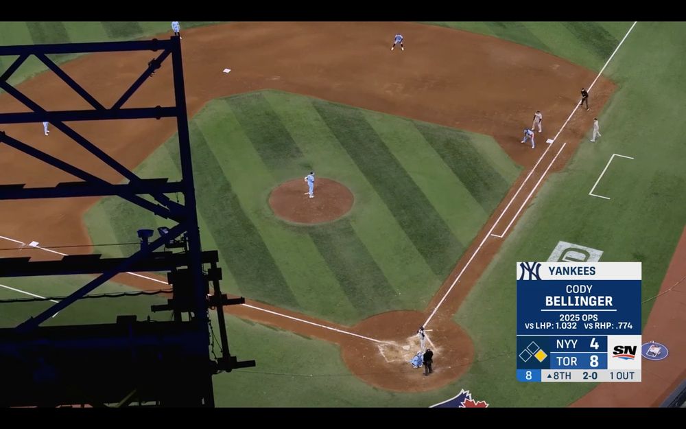 An overhead view of the diamond from the top of the Skydome. The field is brightly lit, and some piece of ceiling machinery makes a jagged-lined dark silhouette on the left third of the screen.