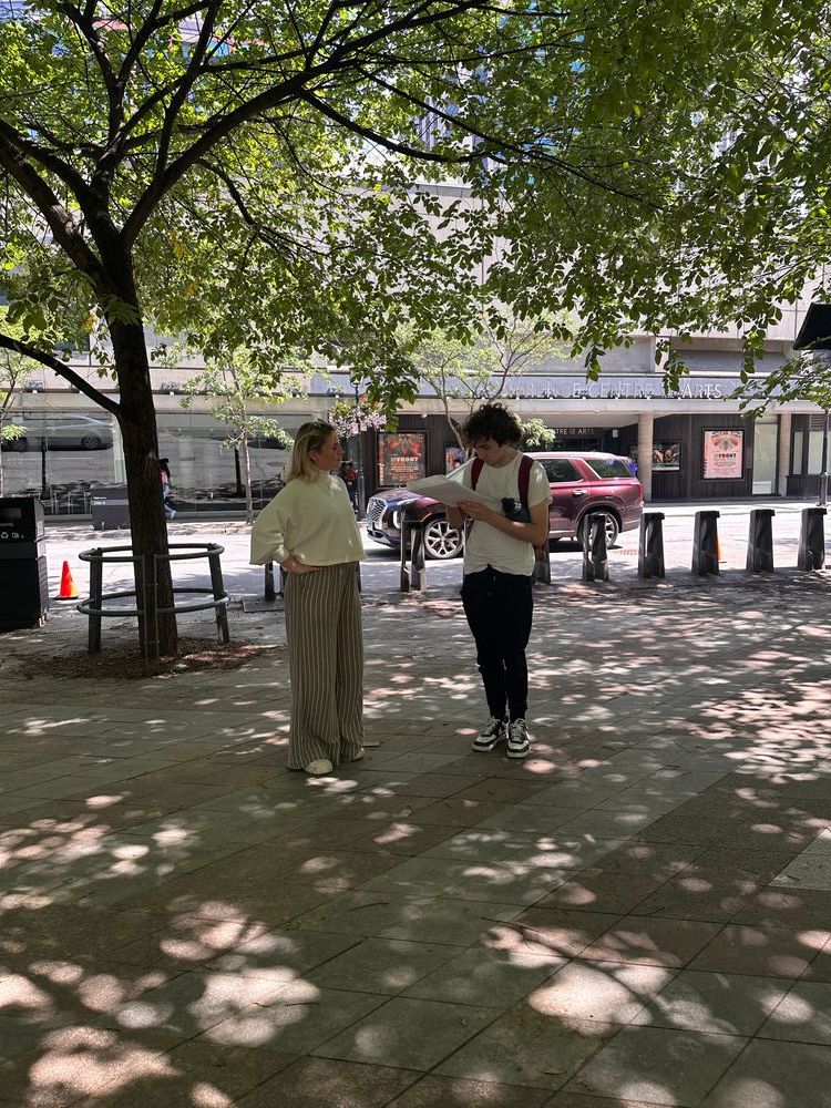 In a park with dappled sunlight, Flo stands next to a man filling in a survey