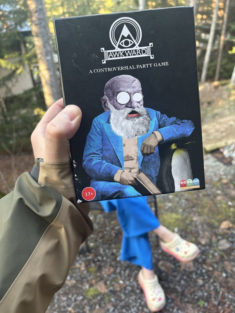Image shows the Awkward card game box with legs matching Wicks coming out from the bottom but wearing crocs