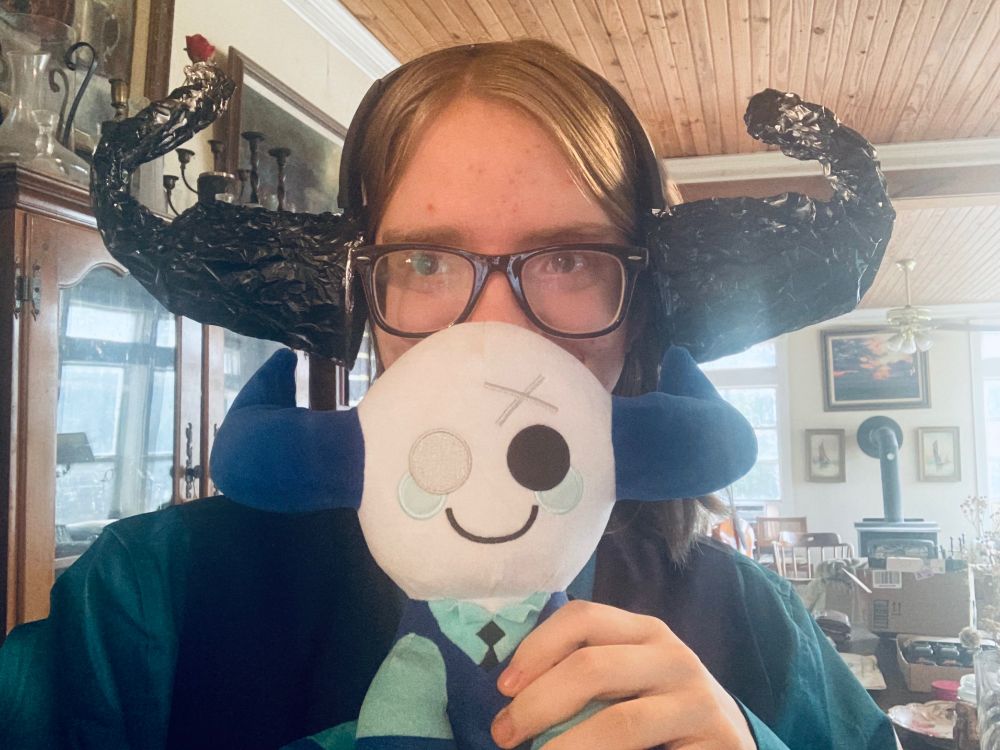 Me wearing a Broker (Phighting) costume with large, twisted horns made from black tinfoil. I am holding a plushie if said Broker (Phighting). I dressed in a dark blue vest with a teal undershirt and a black patterned tie.