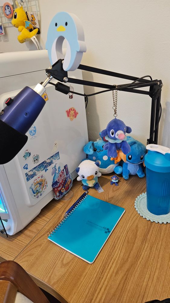 Foto de parte do meu setup, com uma decoração seguindo um tema aquático. Há pelucias de Pokemon (Popplio, Mudkip, Azumarill, Oshawott), e merchs do mesmo tema grudadas ou próximas à case do computador. No apoio de microfone há um Popplio de pelúcia pendurado e um acessório de pinguim encaixado. Por perto tem também um caderno com lapiseira, uma garrafinha e um pequeno sousplat, todos azuis