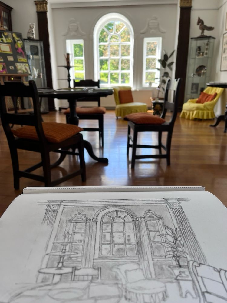 A south facing sitting room in a country house with a large window in three panels overlooking the garden.  The interior space is filled with low chairs, a round table and dining chairs and various stands and ornaments.  At the bottom of the photo is a sketchbook showing a drawing of the view.