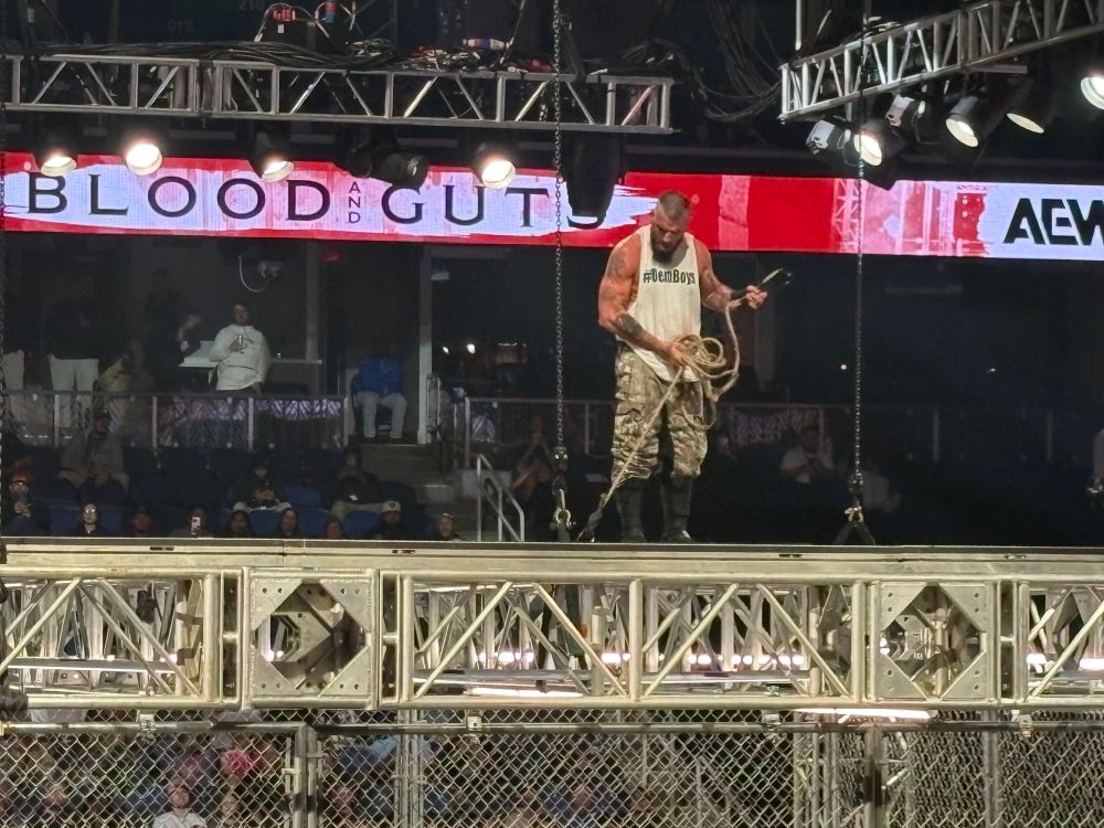 Mark Briscoe of AEW staving on top of the steel cage at Blood and Guts with a hook and a rope before he dropped it down for Kyle O’Reilly to hook half a dozen steel folding chairs to then be winched up by Mark