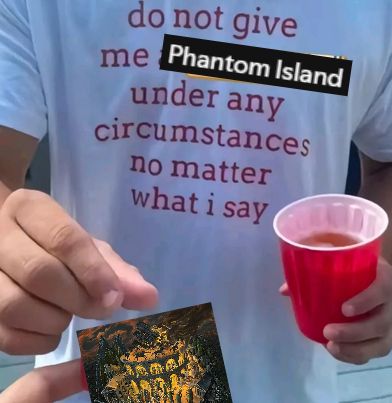 guy wearing shirt that says 'do not give me phantom island under any circumstances no matter what i say' holding a red cup. the person holding the camera is handing the guy phantom island