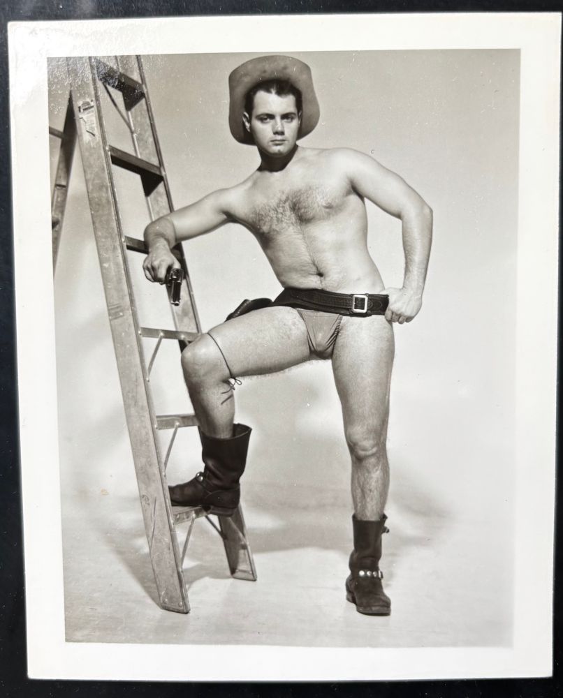 Vintage physique photo of hirsute model wearing cowboy hat, holster, and posing pouch, gun drawn. He leans on a ladder and wears motorcycle boots.
