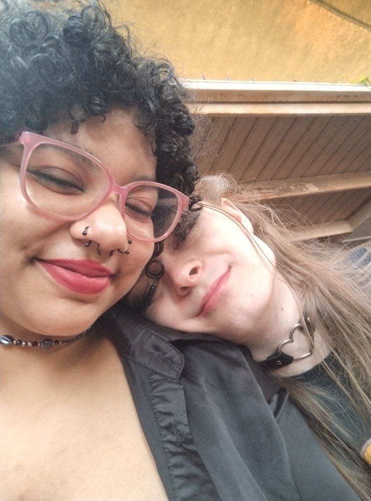 From left to right. It is me with curly black hair smiling at the camera. I have a red lipstick on and pink glasses with a black shirt on. My gf is resting her head on my shoulder with a half smile. She's wearing a faux leather choker with a metal heart in the middle and has long dirty blonde hair