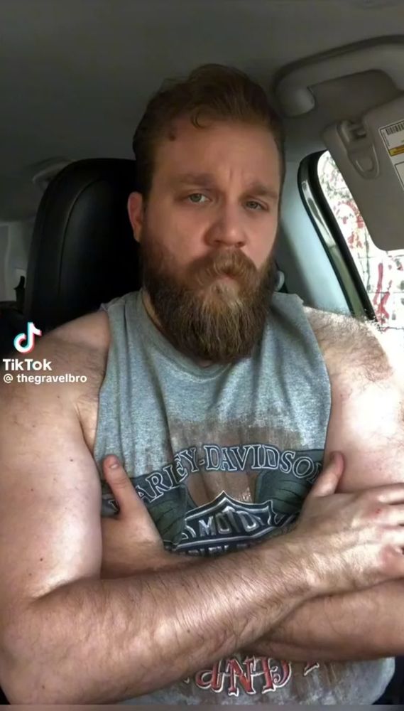 Buff and bearded trans man Leo Macallan, sitting in a car, wearing a sleeveless shirt 