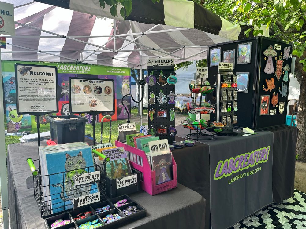 A farmers market setup with a white and black striped tent and bins of cat art prints, sticker displays and black tablecloths. 
