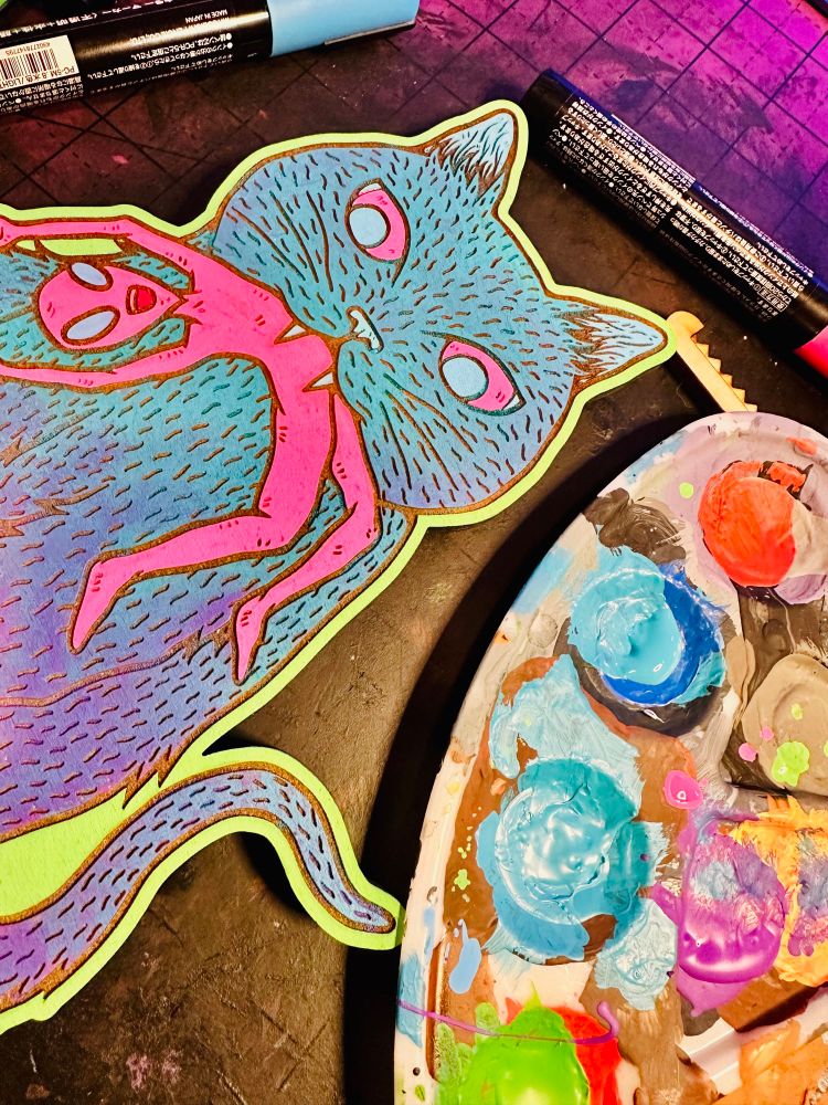 Woodcut cat with a blue and purple gradient with pink and blue eyes with a neon pink alien in their mouth. There is a paint pallette next to it with blue, purple and red paint inside. 