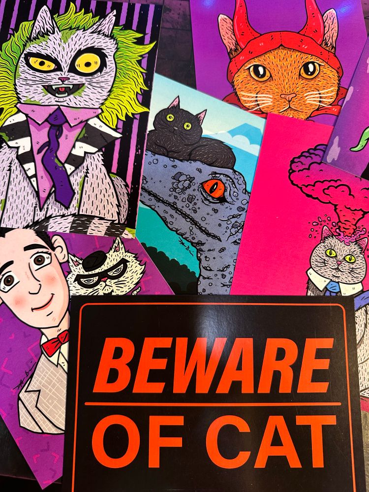 Photo of art prints spread out on a table including a sign that says Beware of Cat with red letters on black background, an illustration of Pee-Wee Herman with Cool Cat, a Raptor with a cat on their head, a cat with a mushroom cloud coming out od the tip of their head, a cat dressed as Beetlejuice, and an orange cat with a devil horn mask. 