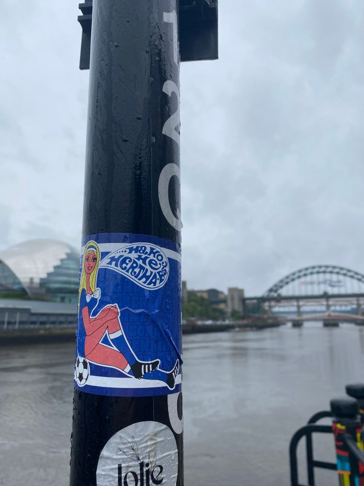 A Hertha BSC sticker on a lamppost in rainy Newcastle, with the Glasshouse and Tyne Bridge in the background. 