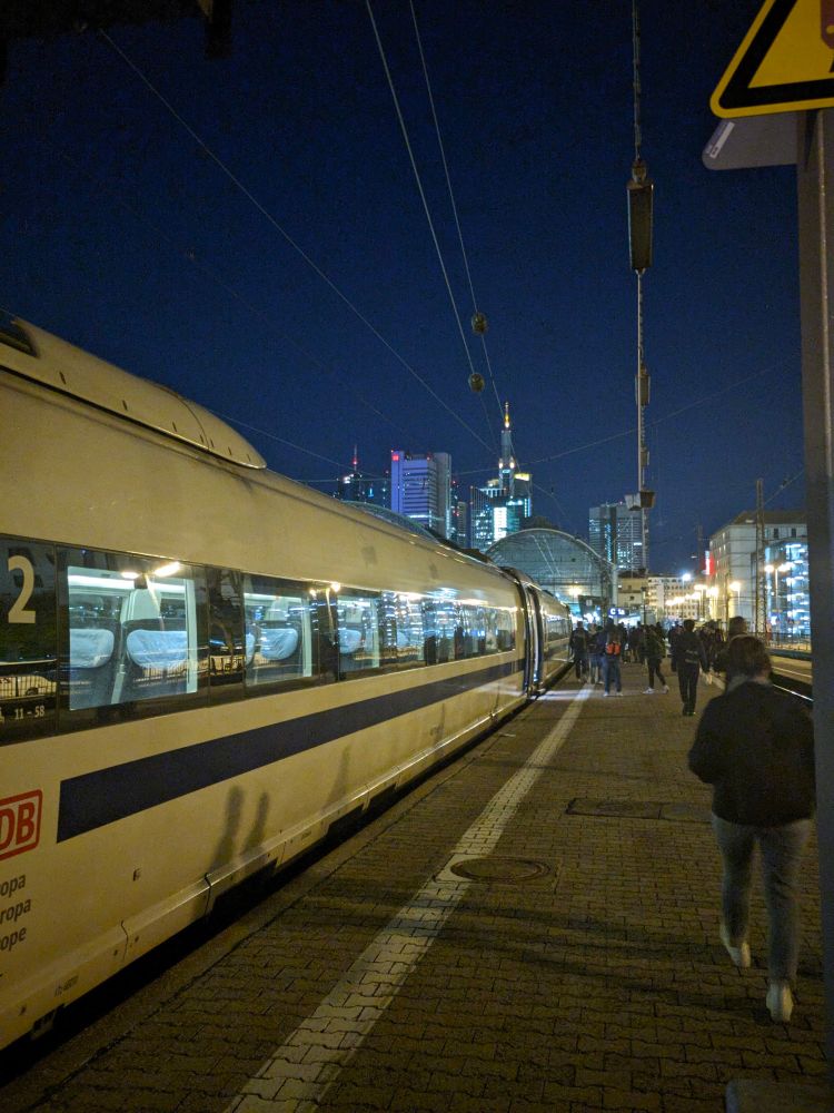 Bahnsteig in Frankfurt Hbf. Am Bahnsteig steht ein Zug der Baureihe 406 mit blauem Zierstreifen, im Hintergrund ist die Frankfurter Skyline zu sehen.
