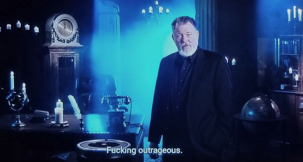 TV's Jonathan Frakes with a white beard and black suit standing in a dark set with candles, the caption text says 