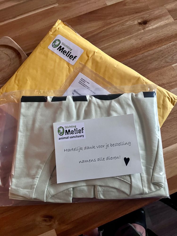 Er ligt een gele enveloppe op een houten tafel, daarop een plastic verpakking met een T-shirt erin, daarbovenop ligt een kaartje met een stempel van stichting Melief en in handschrift gedrukt; “hartelijk dank voor je bestelling namens alle dieren <3 “
